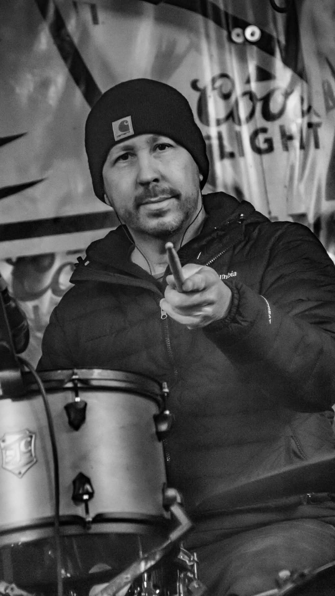 Drummer pointing a stick at the camera from behind the kit — black and white